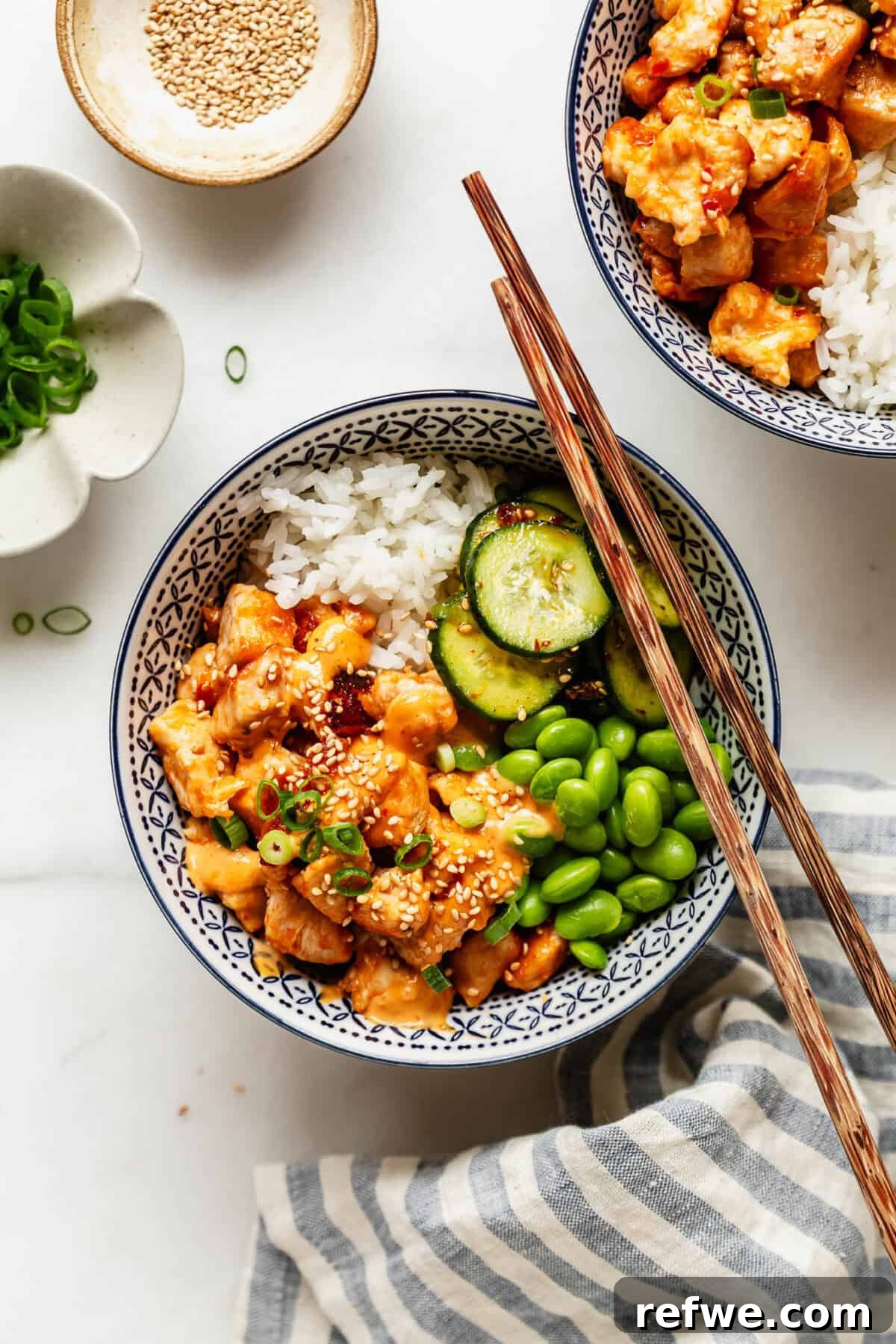 Bang Bang Chicken Bowl Recipe with Spicy Cucumber Salad 10 Two bang bang chicken bowls with rice and vegetables set on a table beside a striped linen napkin.