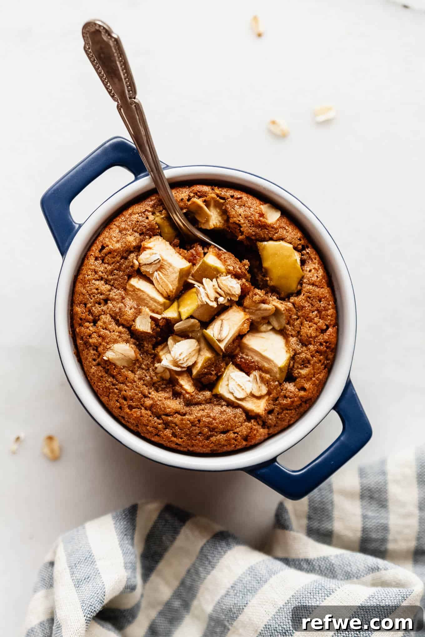 Apple Pie Baked Oats Recipe with Cinnamon 2 A single-serving apple baked oatmeal in a small blue dish topped with fresh apple slices and a spoon.