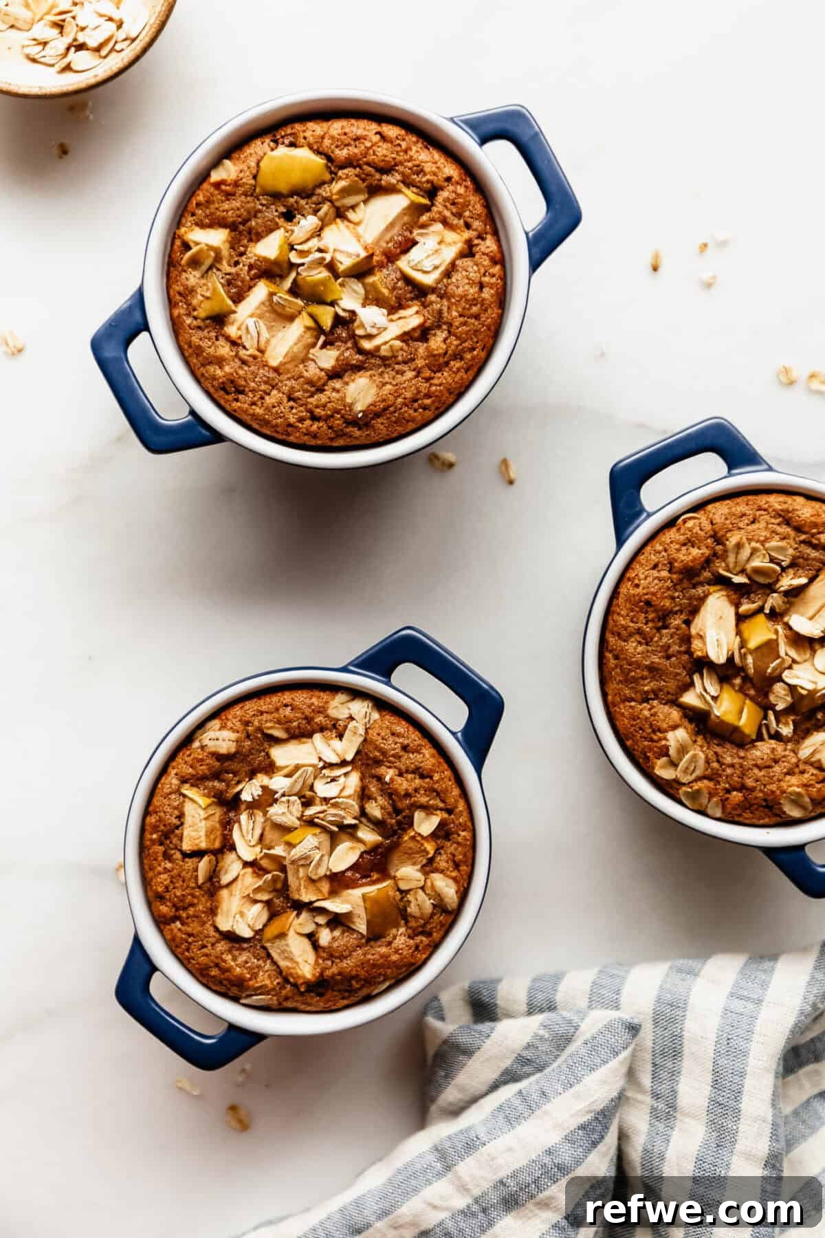 Apple Pie Baked Oats Recipe with Cinnamon 8 Three apple baked oat ramekins staggered on a marble counter beside a striped linen napkin.