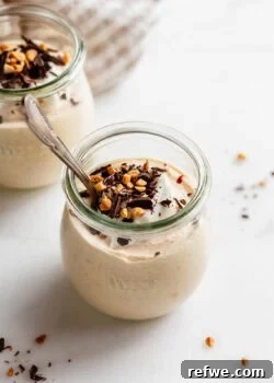 Jar of Greek yogurt peanut butter mousse with a spoon inside, topped with nuts and chocolate shavings.