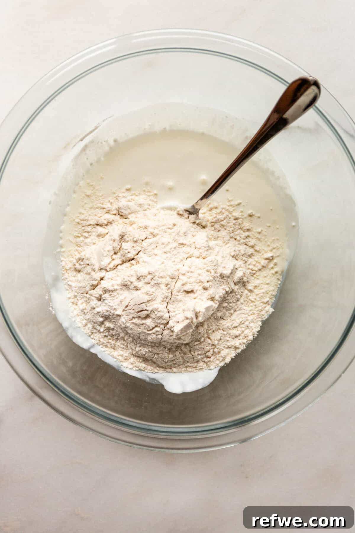 Baked Cottage Cheese Donut Holes Recipe 5 A glass bowl of flour with cottage cheese being stirred together with a spoon to form dough.