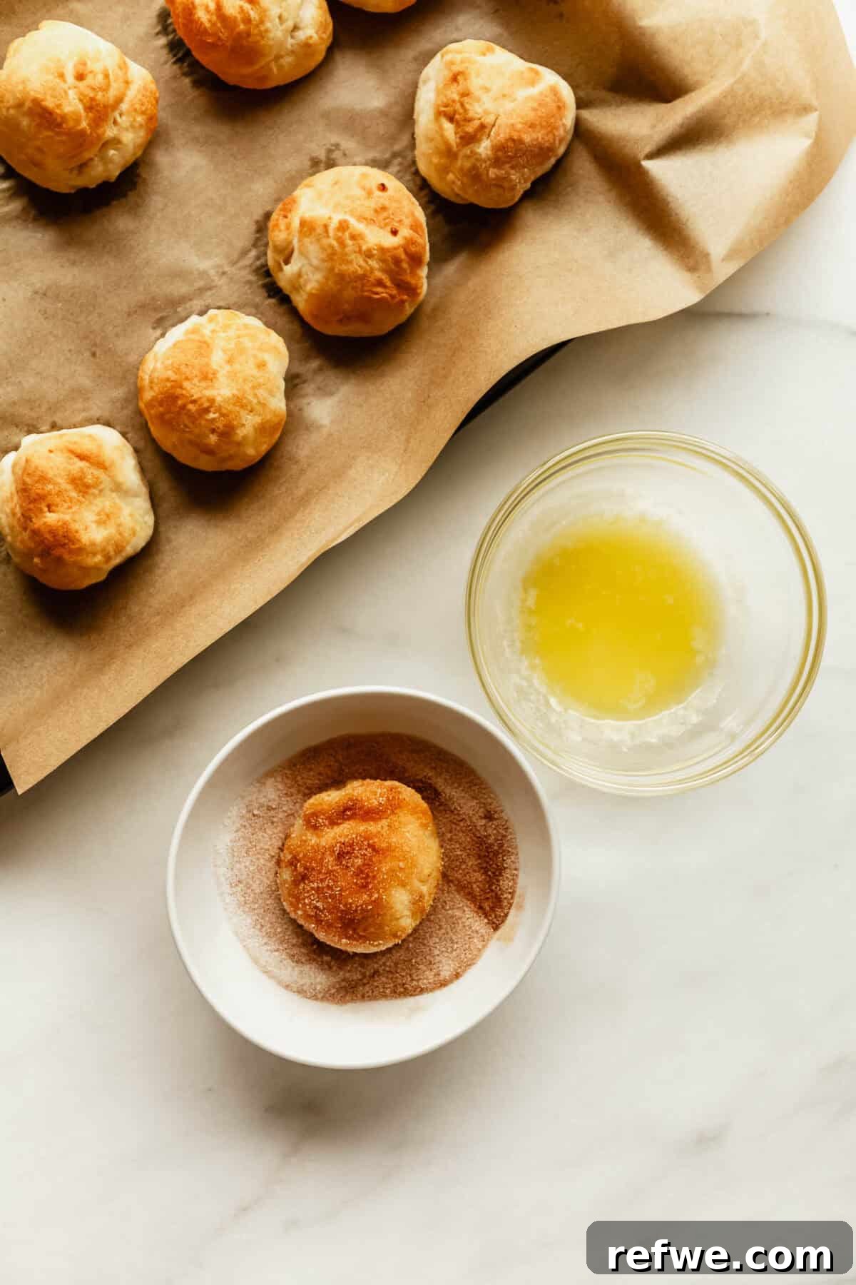 Baked Cottage Cheese Donut Holes Recipe 10 Baked donut holes on parchment paper next to a bowl of melted butter and a bowl of cinnamon sugar with one donut hole being coated.