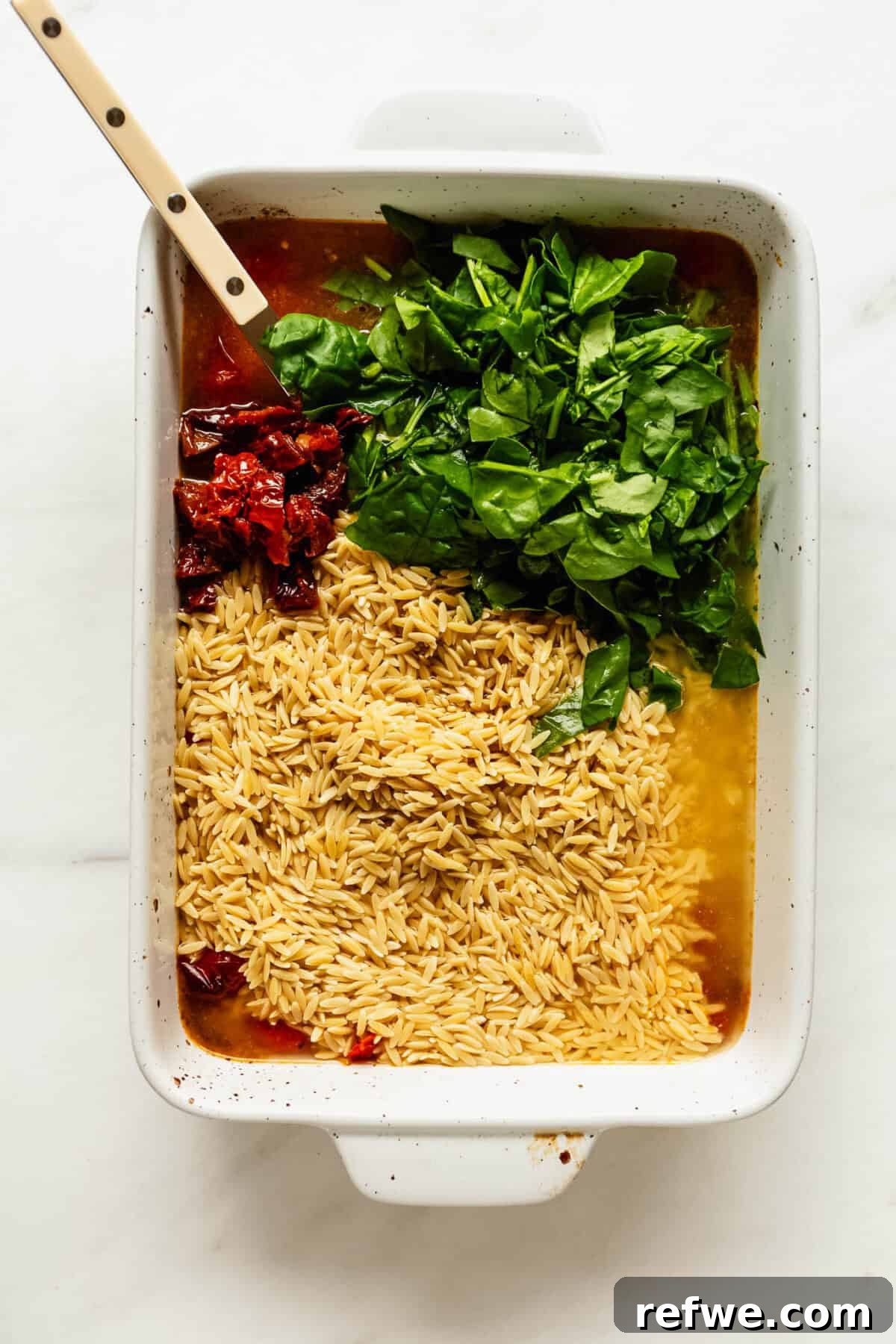 Creamy Baked Feta Orzo with Cherry Tomatoes and Spinach 7 Baking dish of orzo mixed with spinach and sun-dried tomatoes being stirred with a wooden spoon.