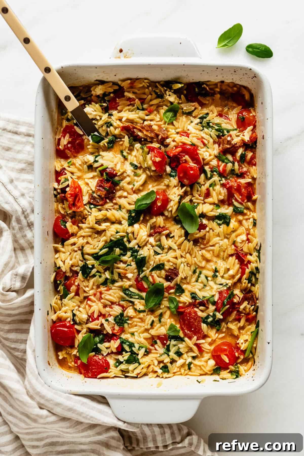 Creamy Baked Feta Orzo with Cherry Tomatoes and Spinach 8 Baked feta tomato orzo garnished with fresh basil leaves in a white baking dish.