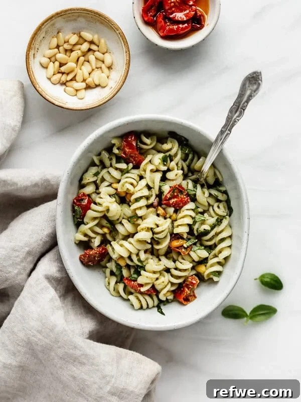 a bowl of pesto pasta salad with a napkin and some pine nuts on the side
